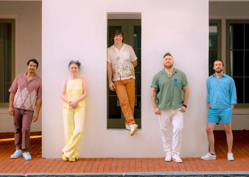 Five people stood against a white wall wearing brightly coloured clothesx