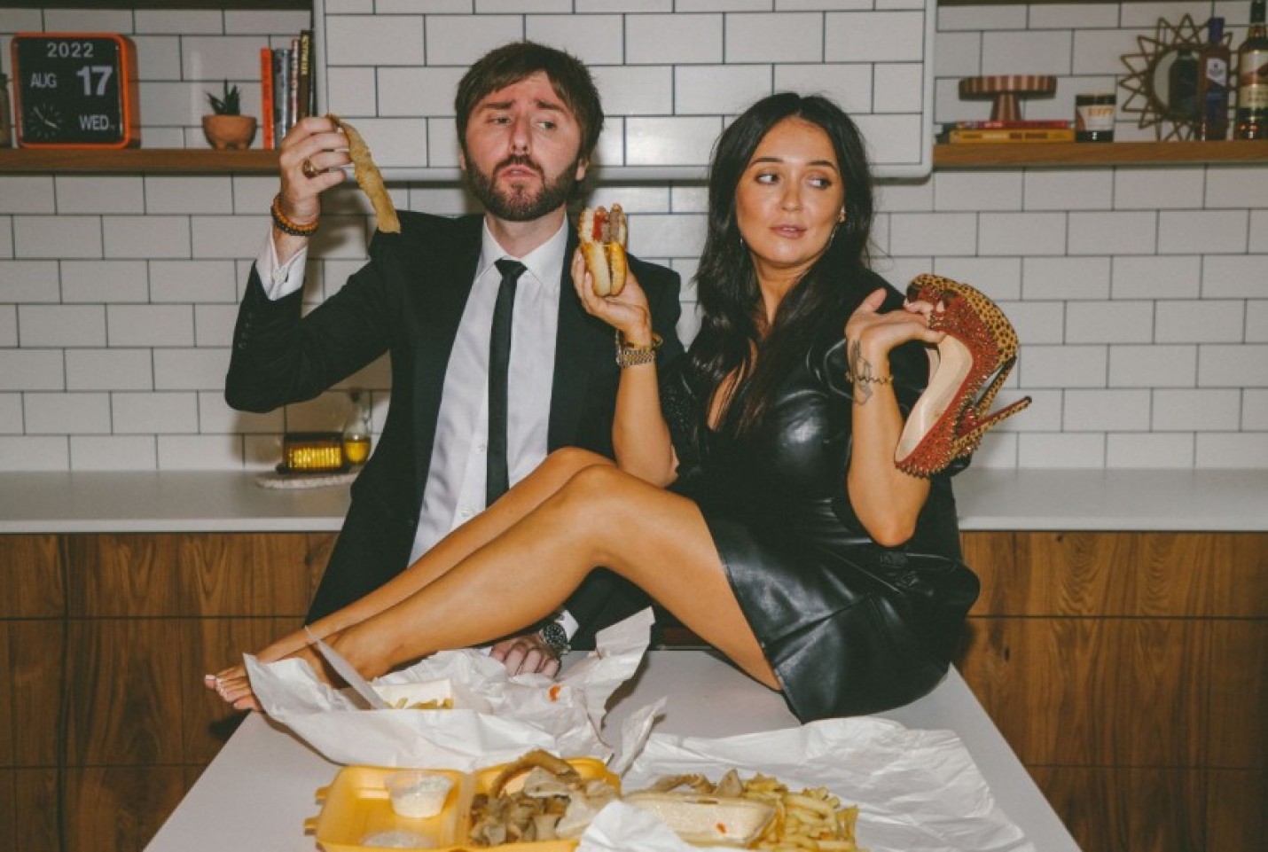 James Buckley standing behind Clair Buckley who is sitting on the kitchen island and both holding food.