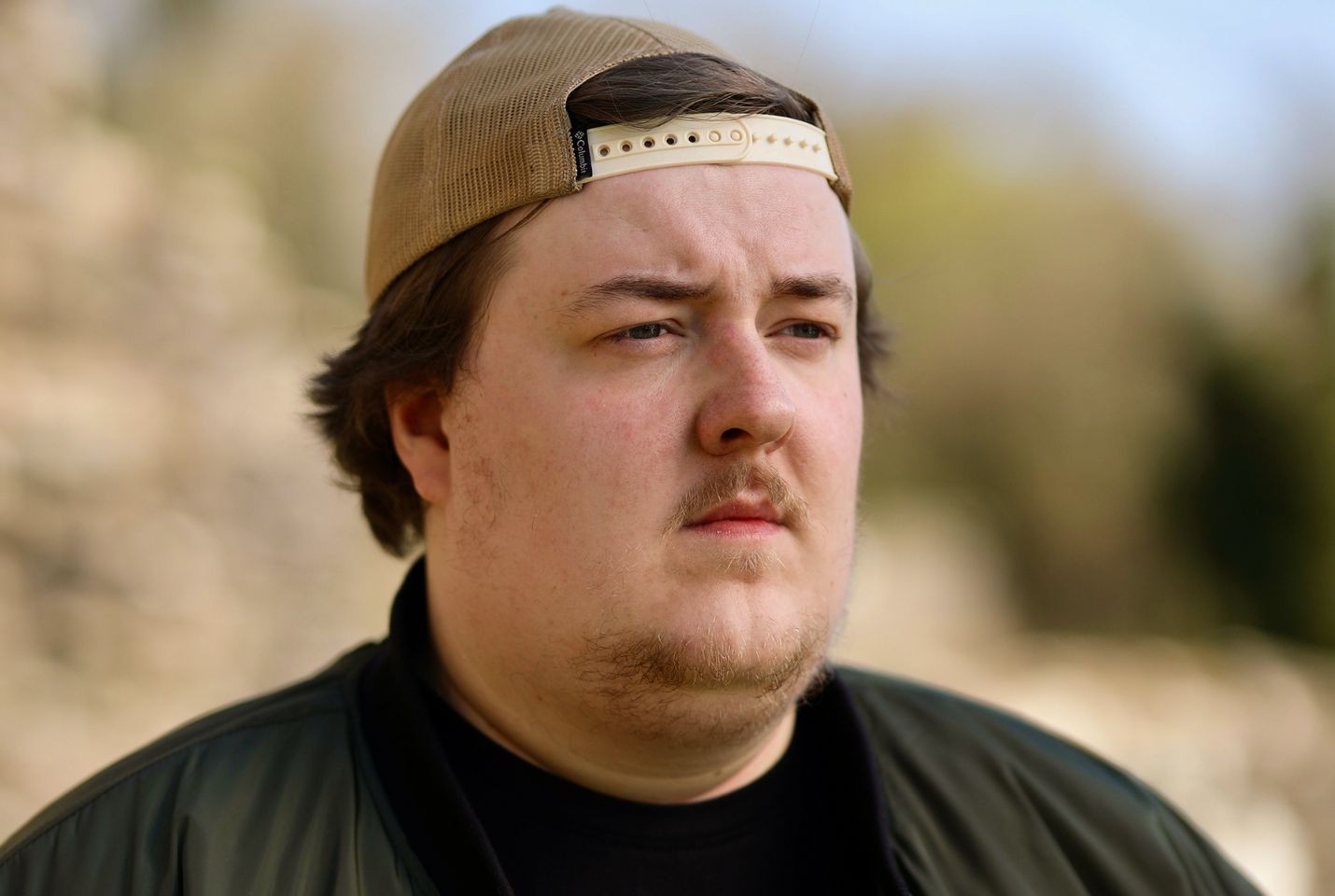A man wearing a backward cap and jacket looks off into the distance