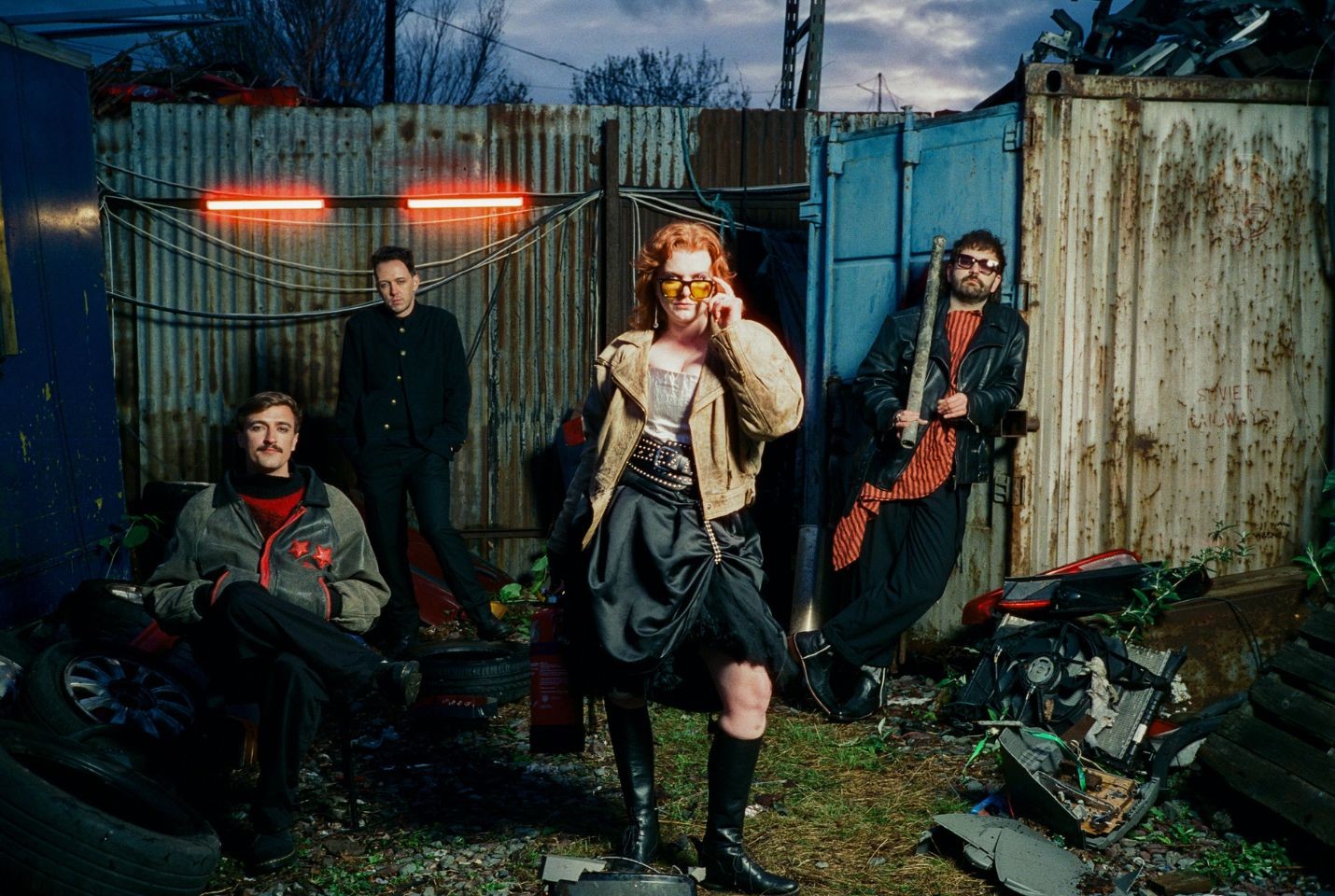 Four band members pose in an industrial yard with corrugated metal walls, scrap metal, and tires. The central figure, a woman with red hair wearing sunglasses, a leather jacket, and a black dress, stands at the front. The other members are dressed in dar