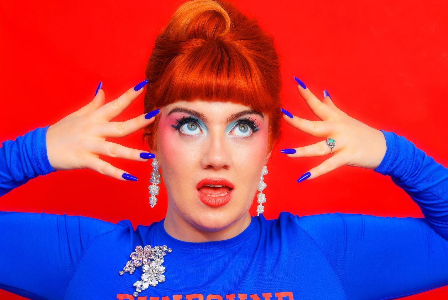 Person with red hair and bold blue makeup poses with hands near face against a red background.