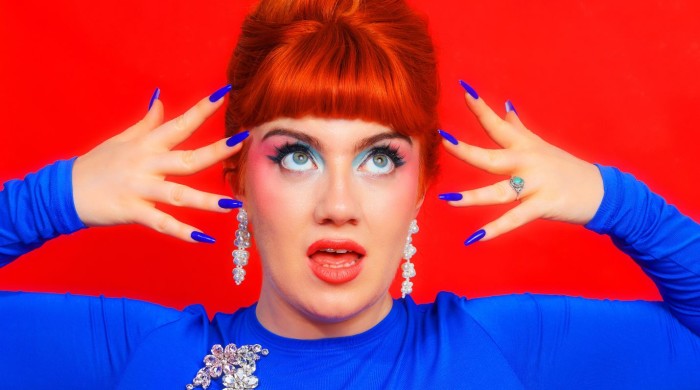 Person with red hair and bold blue makeup poses with hands near face against a red background.