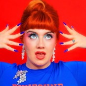 Person with red hair and bold blue makeup poses with hands near face against a red background.
