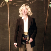 Girl with blonde short, curly hair, standing against brown background