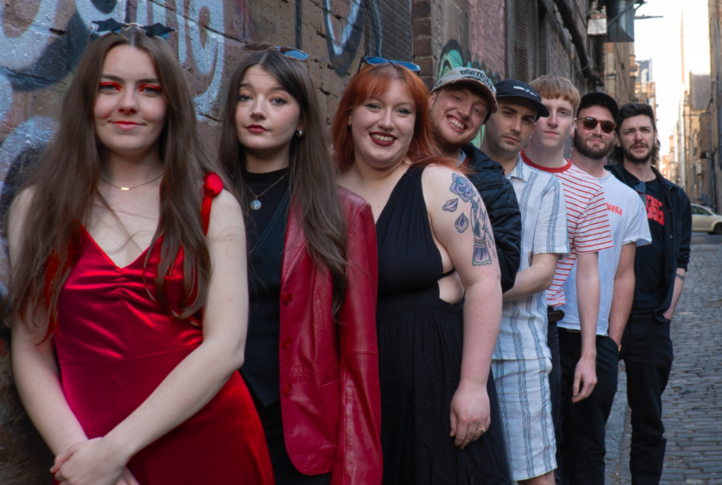 The band Frank's House, with the members Jamie Welsh, Erin Hephzibah, Jennifer Gibson, Katt Kerr, Craig Goodwin, Sam Braes, Finnie Welsh and Stuart Mclntyre queuing up in the street.