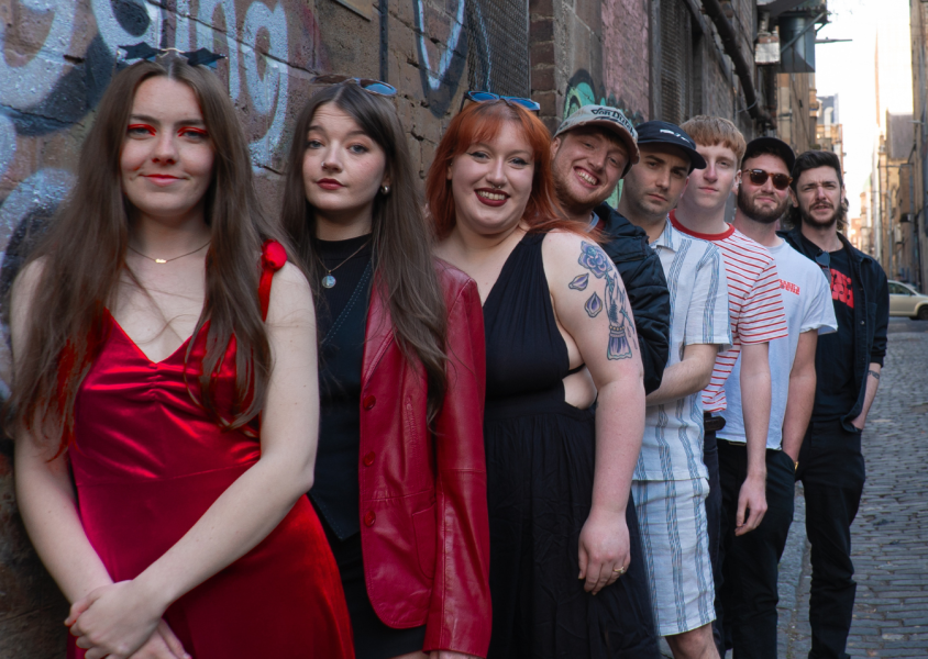 The band Frank's House, with the members Jamie Welsh, Erin Hephzibah, Jennifer Gibson, Katt Kerr, Craig Goodwin, Sam Braes, Finnie Welsh and Stuart Mclntyre queuing up in the street.x