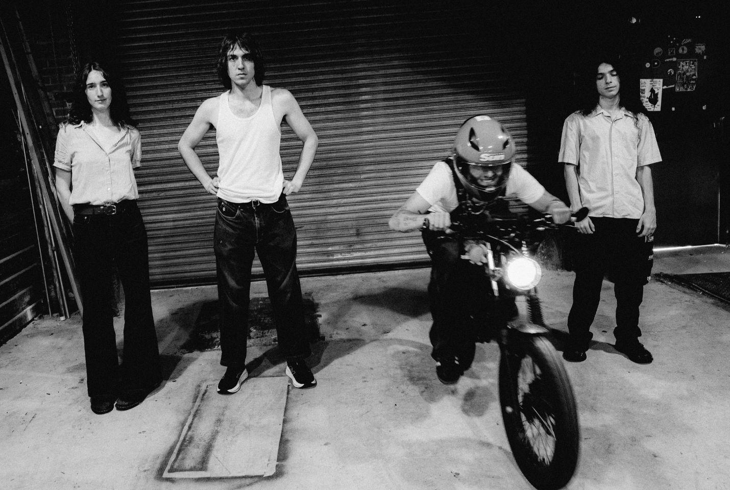 A black and white image of four people in a garage, one is on a motorbike, the rest stood on either side of him.
