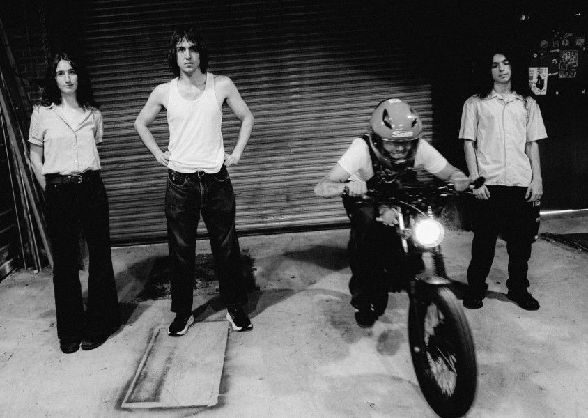 A black and white image of four people in a garage, one is on a motorbike, the rest stood on either side of him. x