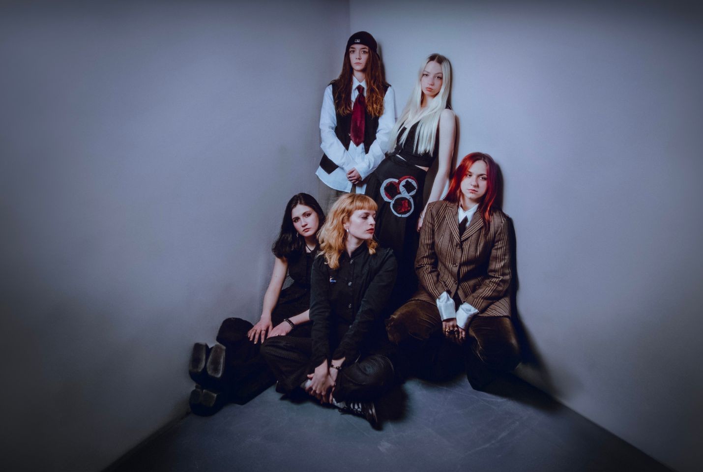 Five women pose together in a minimalist corner of a room with plain gray walls. Two stand at the back, wearing button-up shirts and ties, one with long dark hair under a cap and the other with long blonde hair. Three sit on the floor at the front, dress
