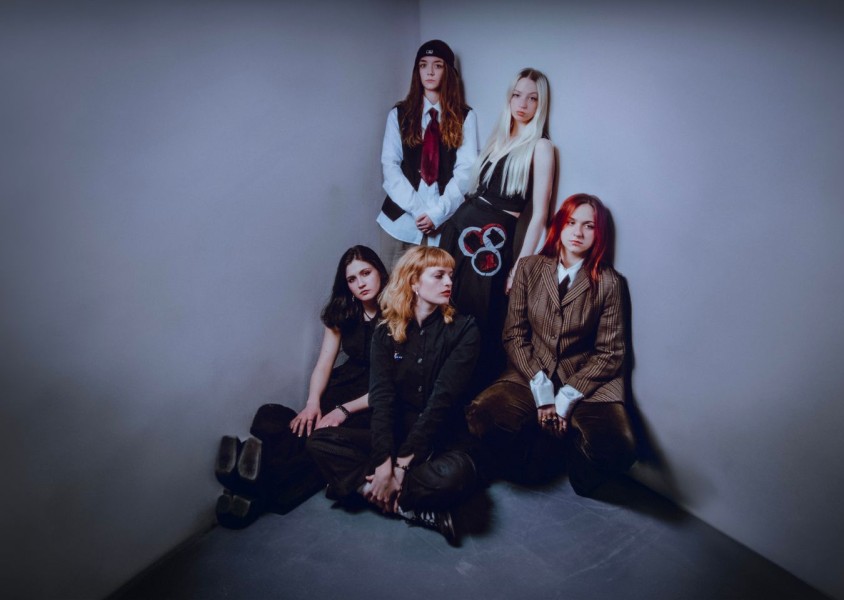Five women pose together in a minimalist corner of a room with plain gray walls. Two stand at the back, wearing button-up shirts and ties, one with long dark hair under a cap and the other with long blonde hair. Three sit on the floor at the front, dressx