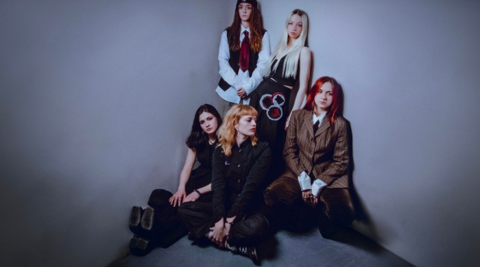 Five women pose together in a minimalist corner of a room with plain gray walls. Two stand at the back, wearing button-up shirts and ties, one with long dark hair under a cap and the other with long blonde hair. Three sit on the floor at the front, dress