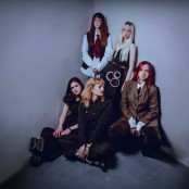Five women pose together in a minimalist corner of a room with plain gray walls. Two stand at the back, wearing button-up shirts and ties, one with long dark hair under a cap and the other with long blonde hair. Three sit on the floor at the front, dress