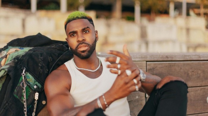 Jason Derulo sitting with a wooden wall behind him, looking into the distance.