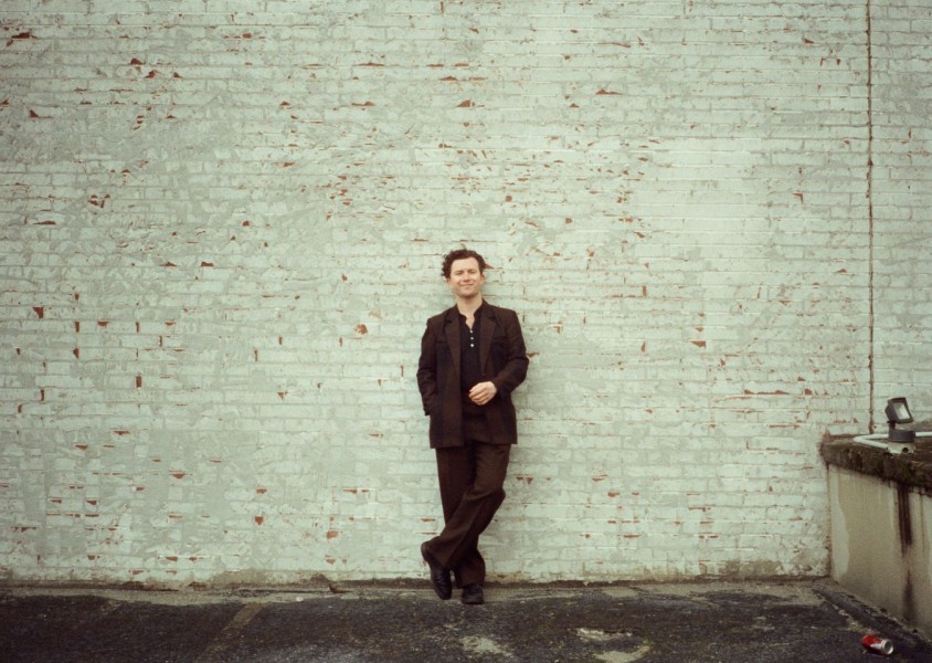 MT Jones smiling into the camera while leaning against a white brick wall.x