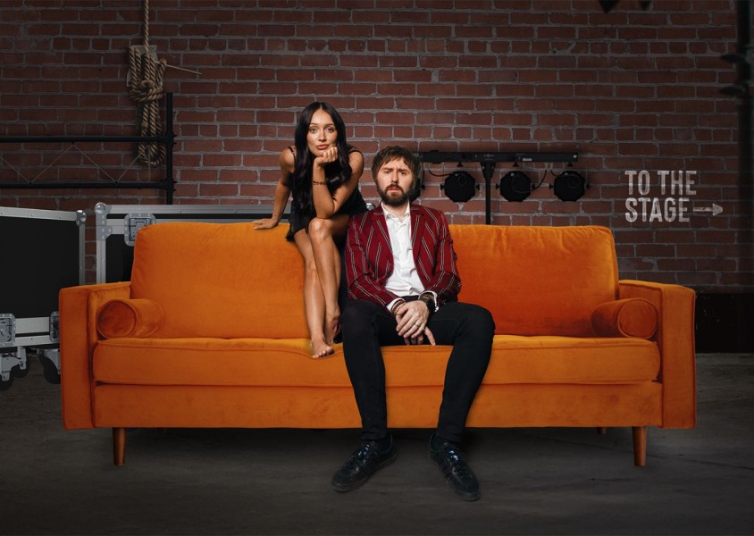 A woman and man sat backstage on an orange sofax
