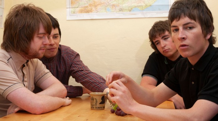 Four young men are sitting around a wooden table in a casual setting. One man in a black polo shirt sits at the front, holding a bunch of grapes, while the others sit close together, looking at the table. A ceramic mug is also on the table. A map is pinn