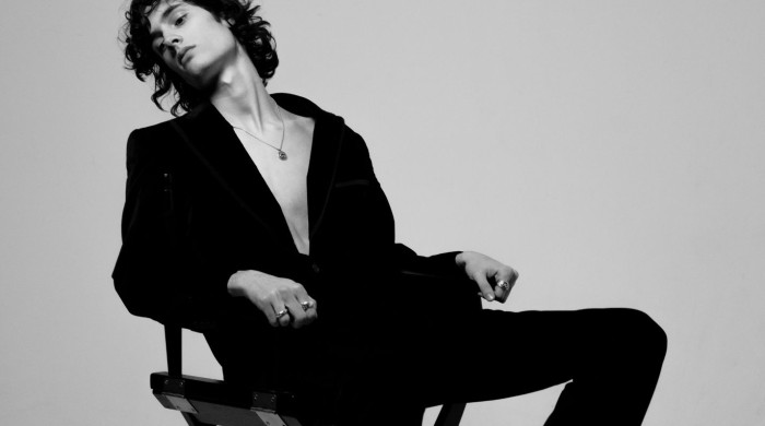 new york based musician sombr sat leaning back on a chair in a black suit with rings and a necklace on