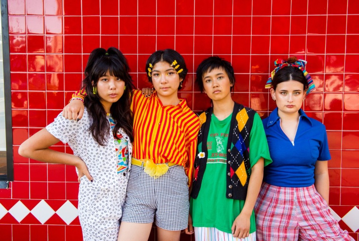The Linda Lindas band, with members leaning against a red brick wall, looking at the camera.
