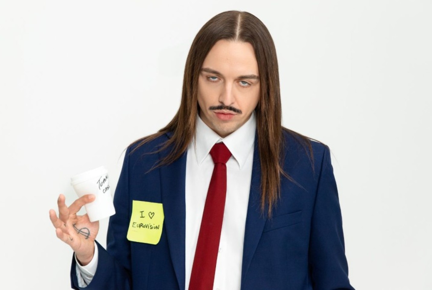 Tommy Cash wearing a blue suit with a yellow posit note on it and holding a cup, while looking at the camera with a straight face.