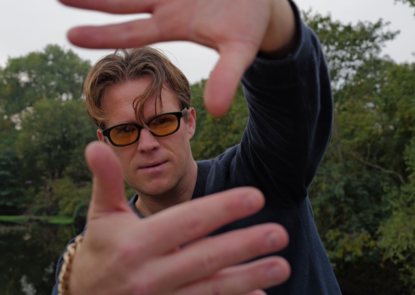 A man with blonde hair wearing orange lensed glasses and a blue jumperx