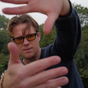 A man with blonde hair wearing orange lensed glasses and a blue jumper