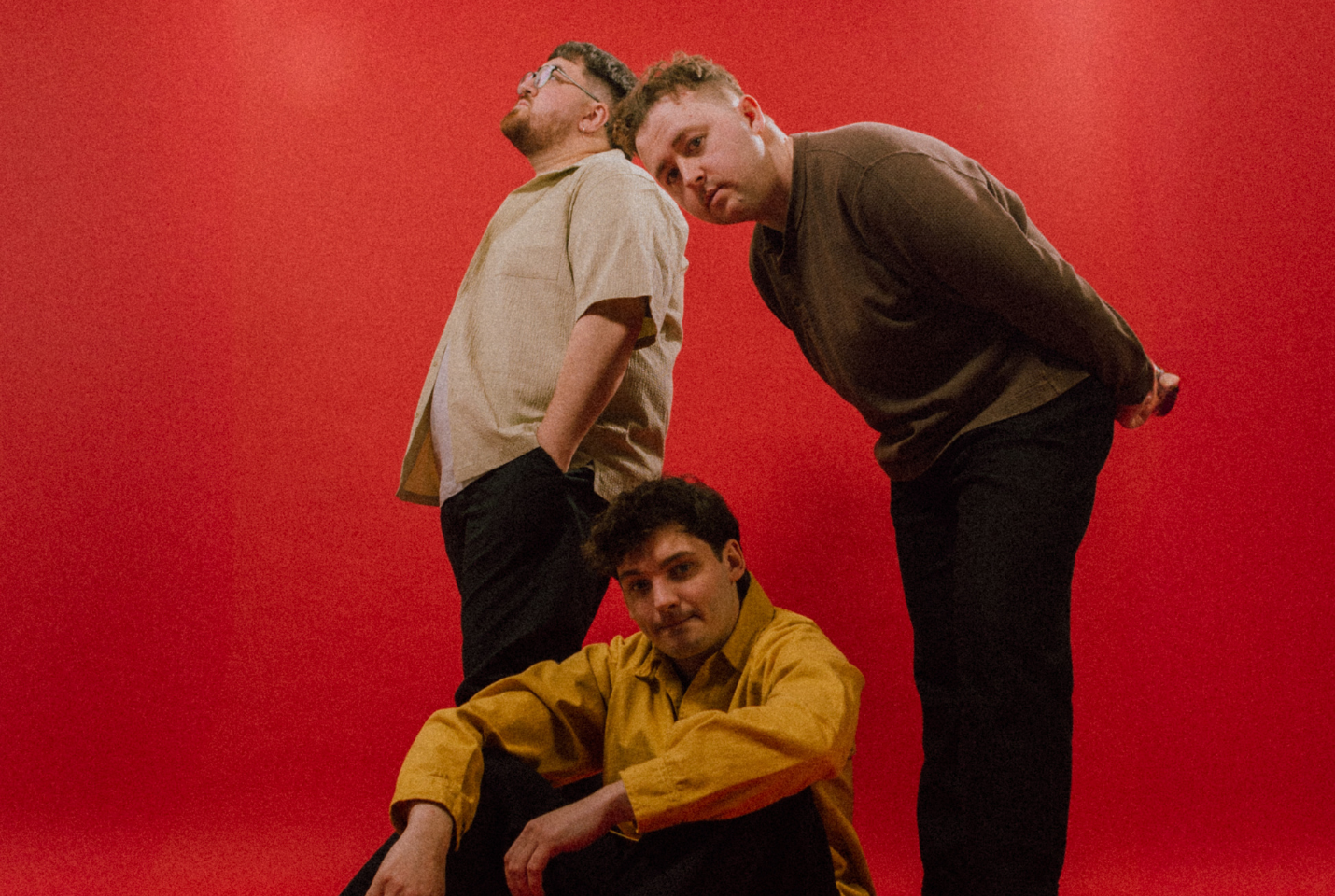 The 3 members of the band 'Vistas' in front on a red backdrop, one member sitting on the floor, another bending over him whilst standing and the third member standing behind looking to something in the top left corner.