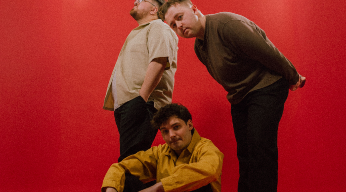 The 3 members of the band 'Vistas' in front on a red backdrop, one member sitting on the floor, another bending over him whilst standing and the third member standing behind looking to something in the top left corner.