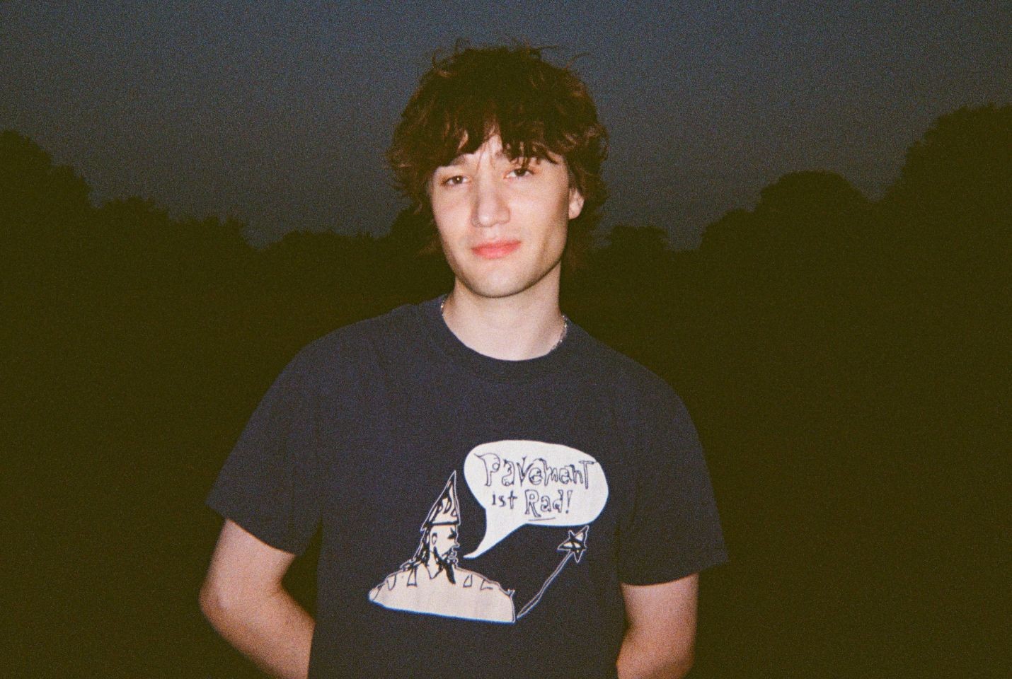 A man with messy brown hair wearing a navy tshirt and silver chain