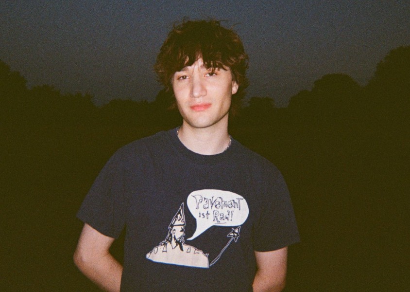 A man with messy brown hair wearing a navy tshirt and silver chainx