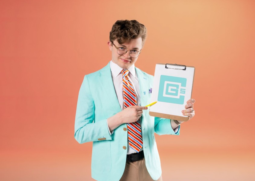 A man wearing a white shirt, brown trouser, a light blue jacket, striped tie and glasses holding a clip board and yellow pen standing in front of an orange backdropx