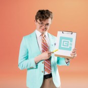A man wearing a white shirt, brown trouser, a light blue jacket, striped tie and glasses holding a clip board and yellow pen standing in front of an orange backdrop